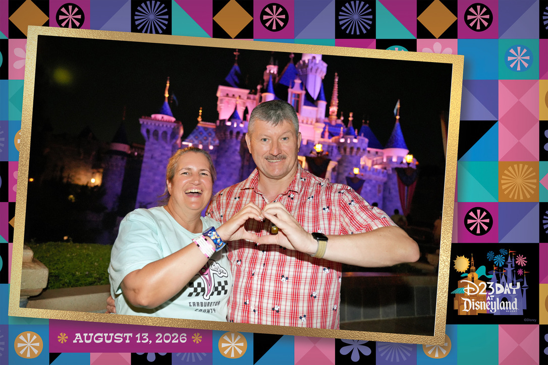 Mausgecheckt mit einem Handherz vor dem beleuchteten Disneyland-Schloss im bunten D23-Day-Bilderrahmen
